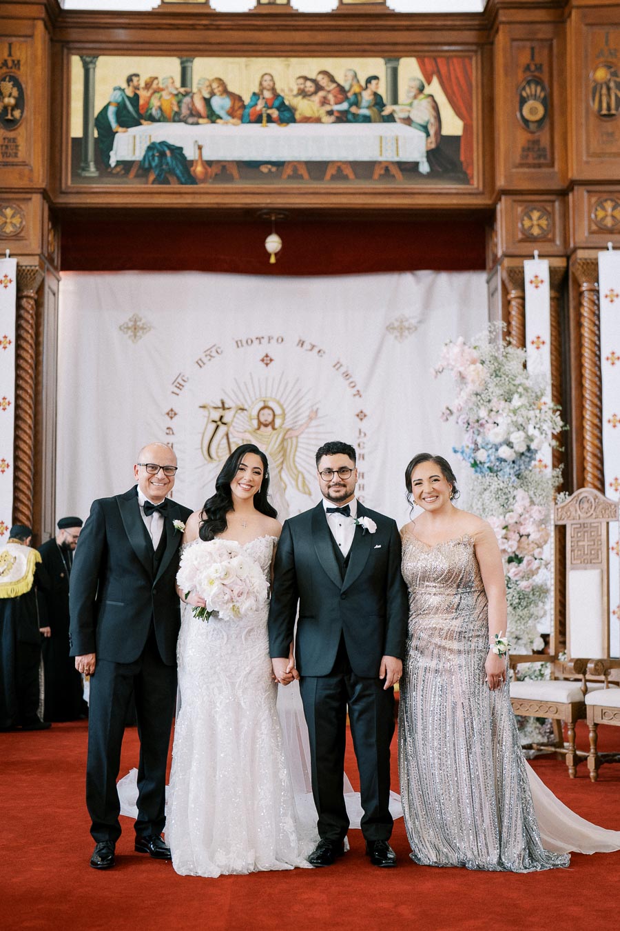 Wedding party posing in ornate church setting, featuring a bride in a lace gown holding a bouquet of white flowers, a groom in a black tuxedo, and well-dressed guests. Beautiful floral arrangements and religious artwork in the background frame the celebration.