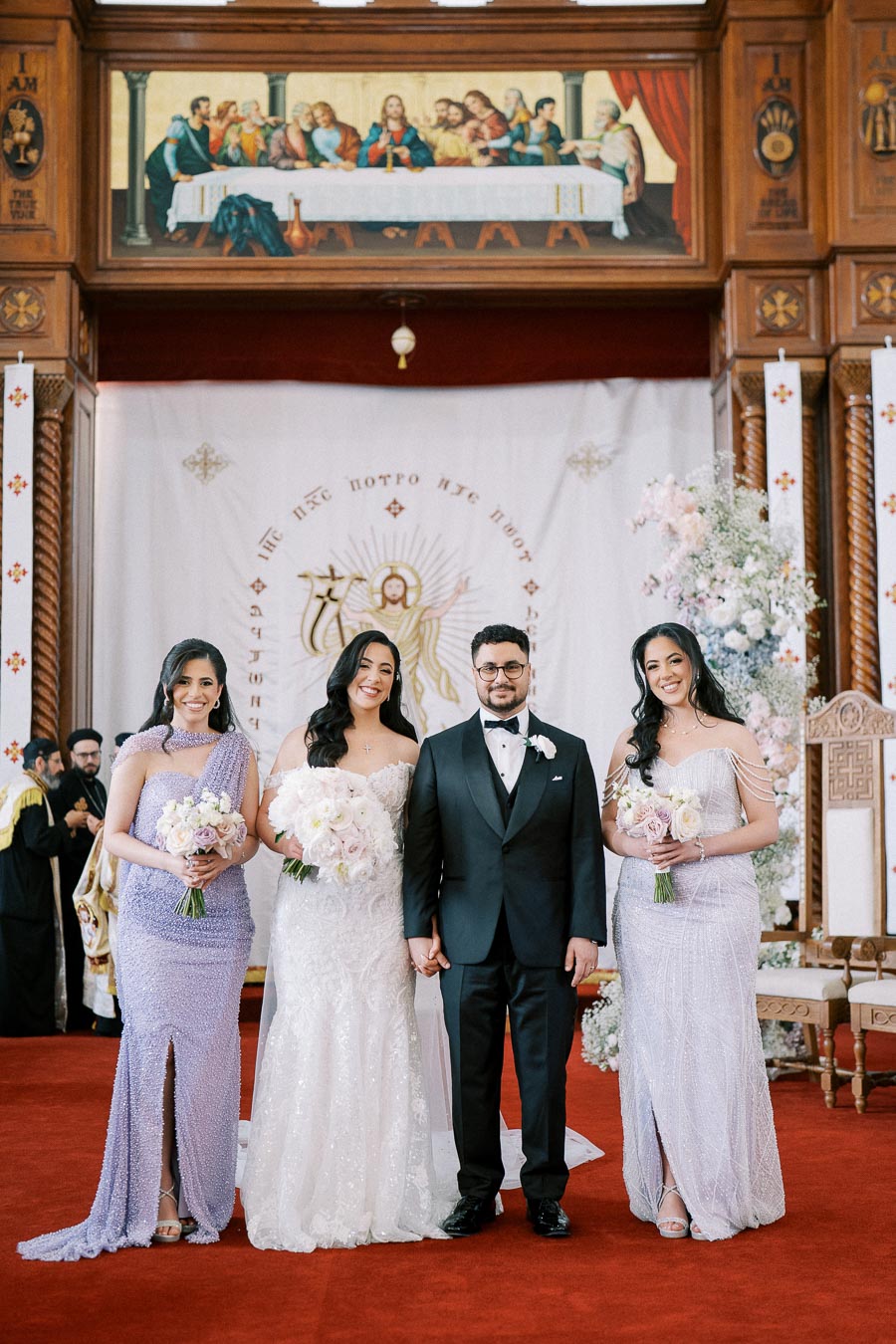 Wedding ceremony in an ornate church, featuring a bride in a white gown and a groom in a black suit with bridesmaids in lilac dresses holding bouquets, under a religious banner and painting.
