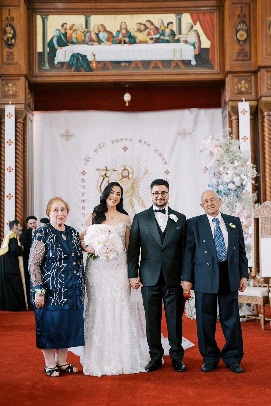 Wedding couple with family members in church setting, featuring elegant traditional attire, floral decorations, and religious iconography in the background.