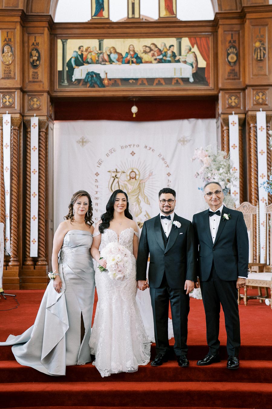 A bride and groom stand with their parents on the steps of a beautifully adorned church altar, featuring intricate wooden decorations and a mural. The bride is in a white lace gown holding a bouquet, while the groom and father are in formal black suits, and the mother is in an elegant silver dress.