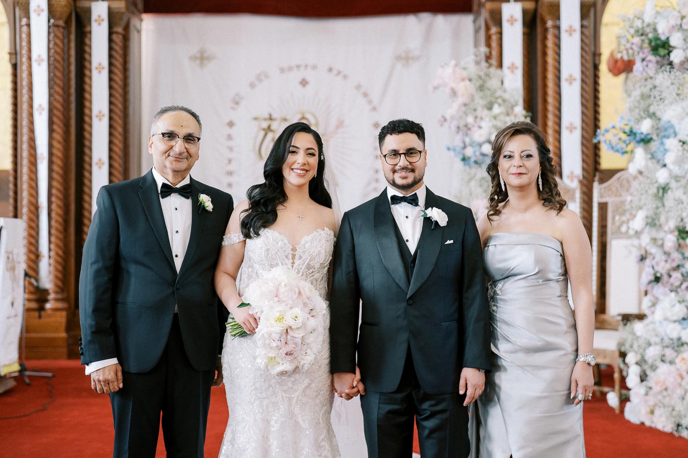 A happy couple stands together with their parents in a beautifully decorated church, celebrating their wedding ceremony. The bride is in a lace gown holding a bouquet of flowers, while the groom and parents are dressed in formal attire.