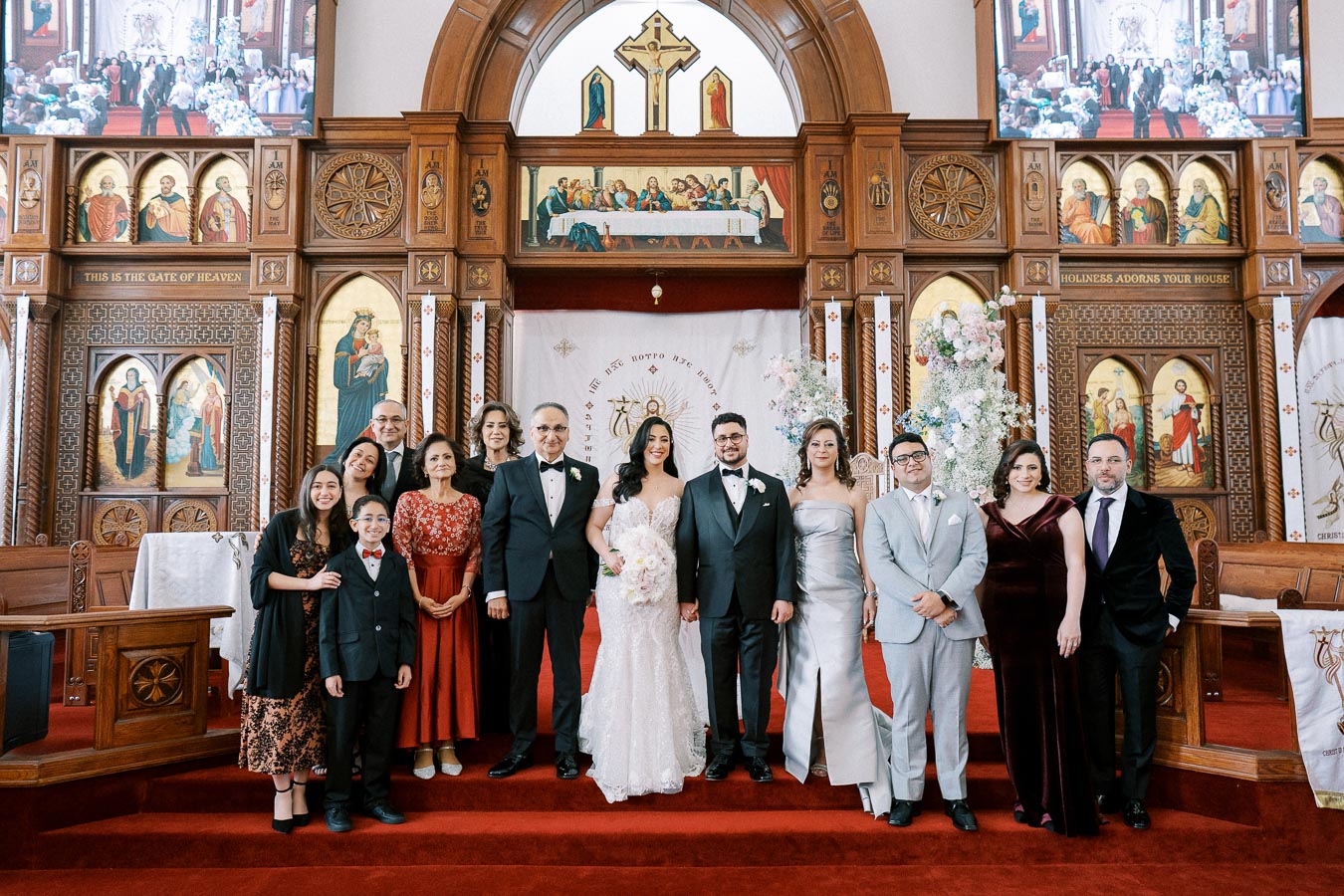 A wedding ceremony group photo inside a beautifully adorned church. The bride and groom stand in the center, surrounded by family members dressed in formal attire. The church features ornate woodwork, vivid religious iconography, and a red carpeted altar area.