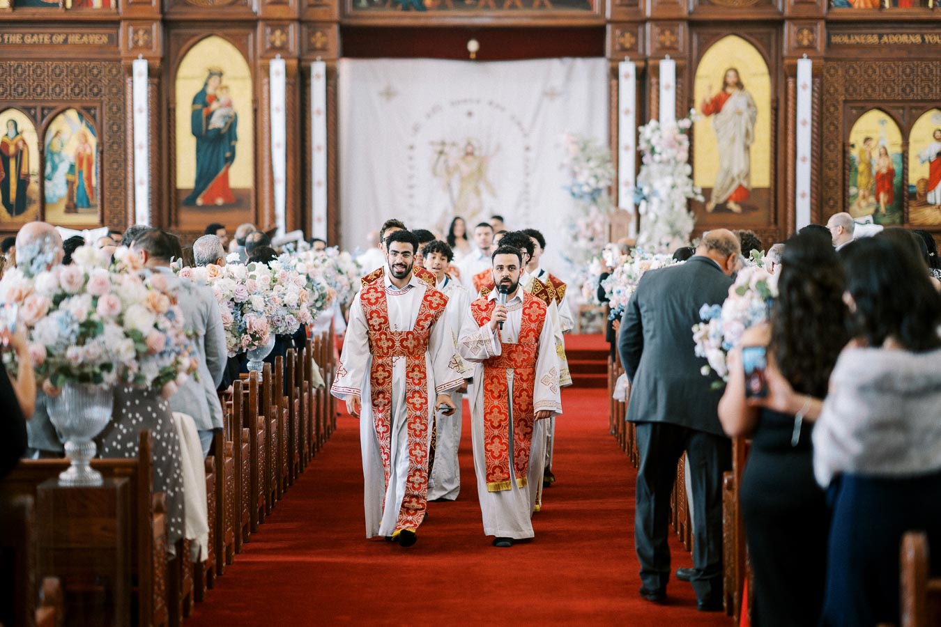 A procession of clergy in white and red vestments walking down the aisle of an ornately decorated church with floral arrangements and religious iconography, during a ceremony.