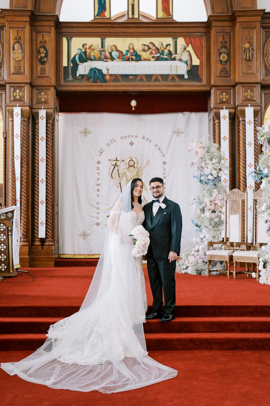 A bride and groom stand smiling on their wedding day within an ornately decorated church. The bride wears a flowing white gown with a long veil, while the groom is dressed in a classic black tuxedo. The backdrop features religious artwork and floral arrangements, creating a beautiful and serene scene.