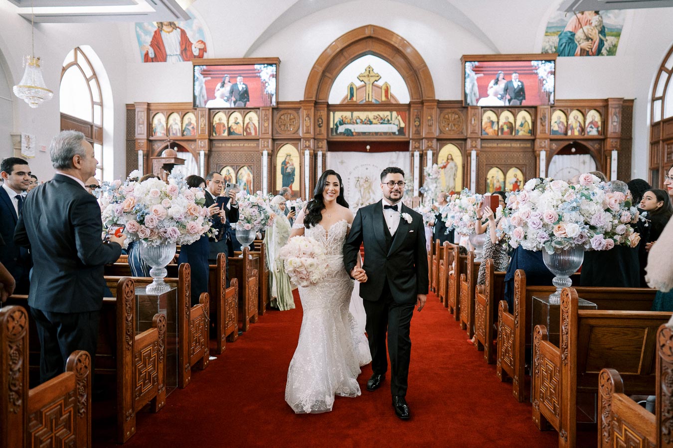 Bride and groom walking down the aisle of a beautifully decorated church, surrounded by guests and pink floral arrangements.