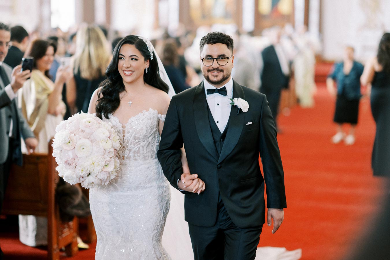 A bride in a lace wedding dress holding a bouquet of white flowers, walking hand-in-hand with a groom in a black tuxedo down the aisle of a church.