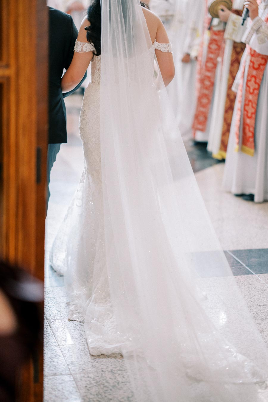 A bride in a white wedding gown with a long train walks down the aisle, accompanied by a person in a suit, during a traditional ceremony with officiants in the background.
