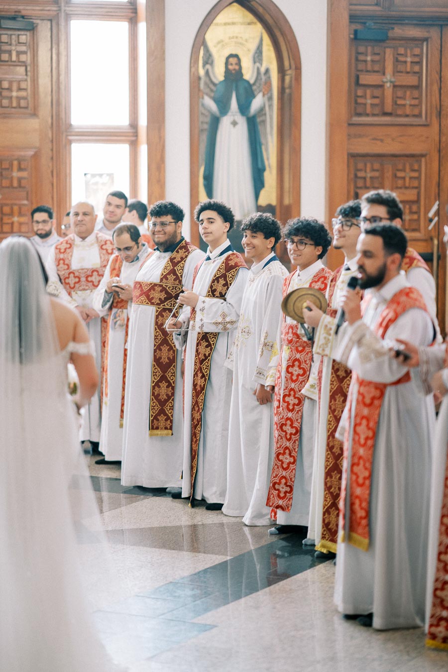A group of clergy in ornate robes participating in a traditional church ceremony, standing in rows inside a church with wood-paneled walls and a religious painting in the background.