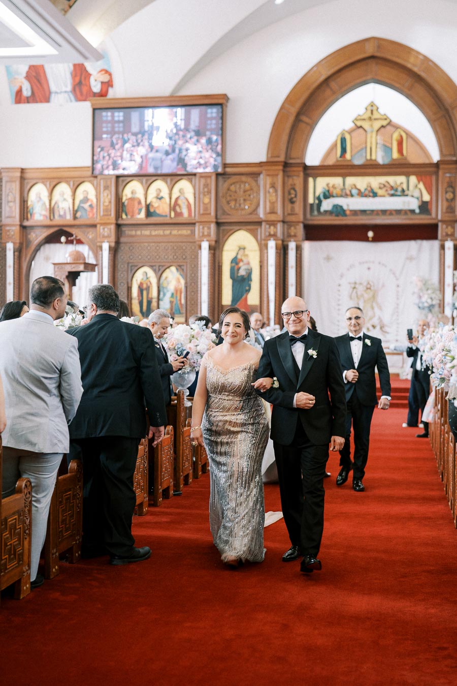 A couple walking down the aisle in a beautifully decorated church during a formal ceremony, with guests on either side and intricate religious icons on the walls.