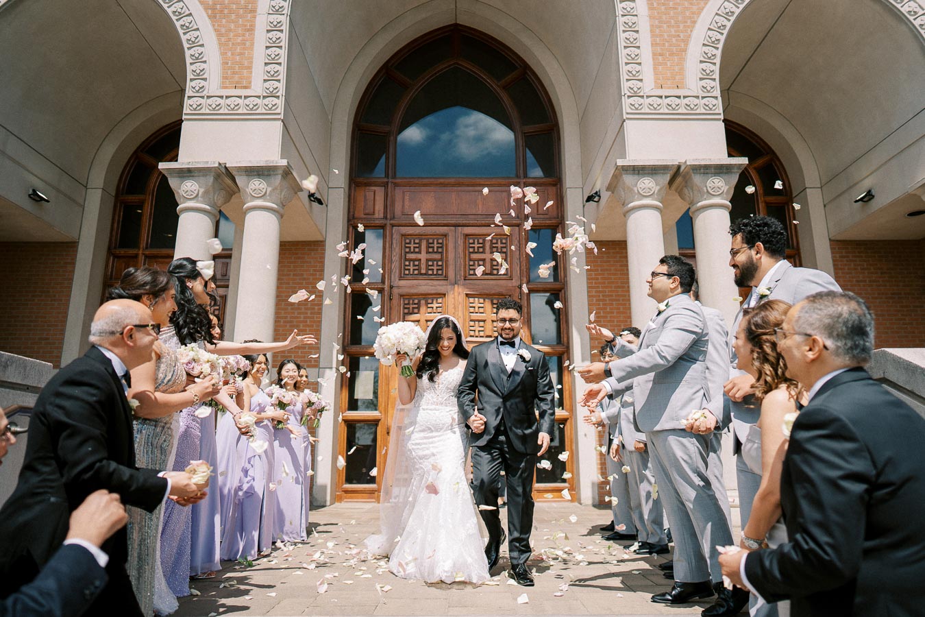 Newlywed couple joyfully exits church amidst a shower of rose petals, surrounded by happy wedding guests dressed in formal attire.