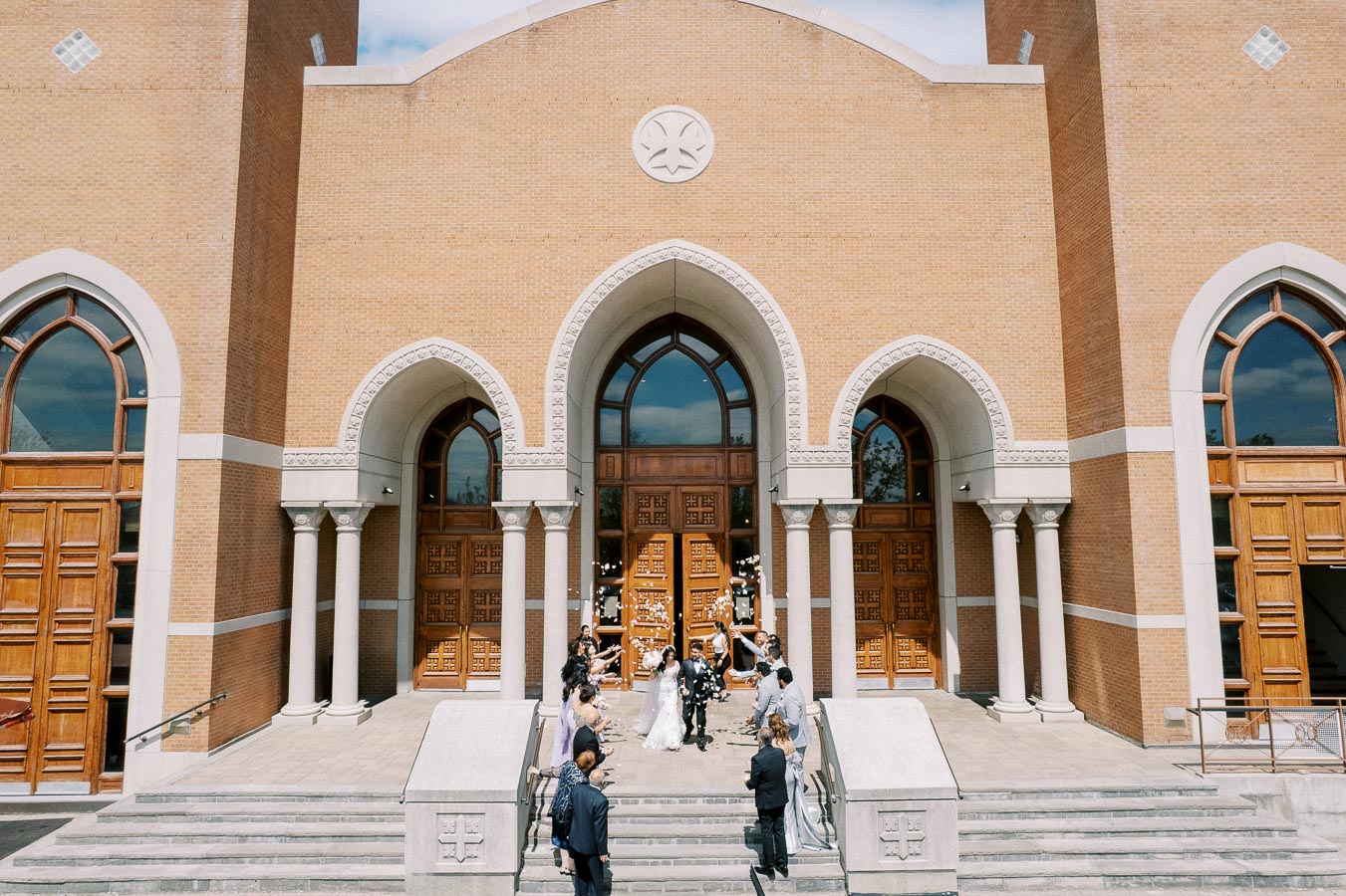Wedding celebration outside a large church with arched wooden doors, as the newlyweds are greeted by guests on the steps.