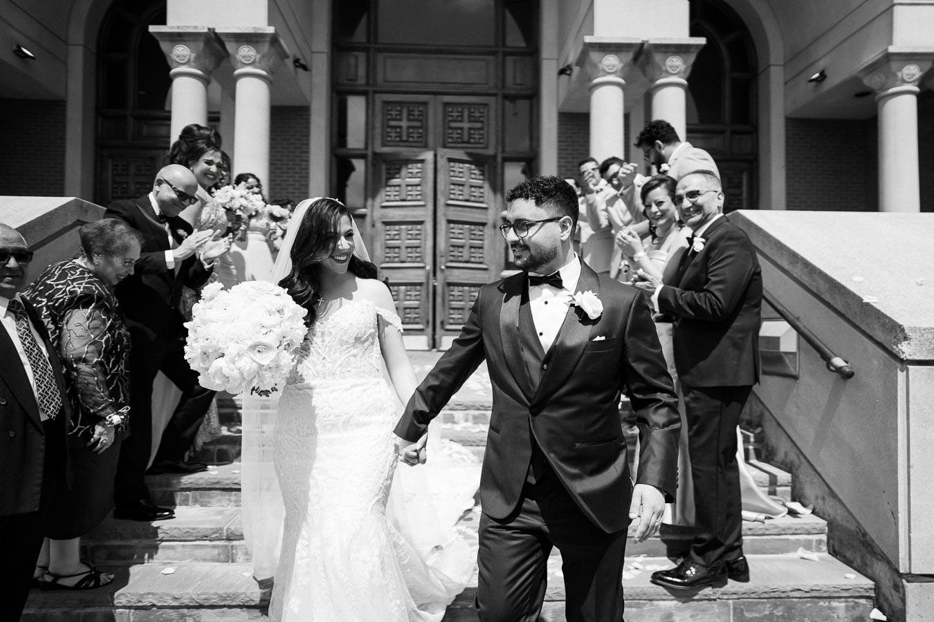 Wedding couple holding hands and smiling as they exit a building, surrounded by happy guests clapping and celebrating on the steps.