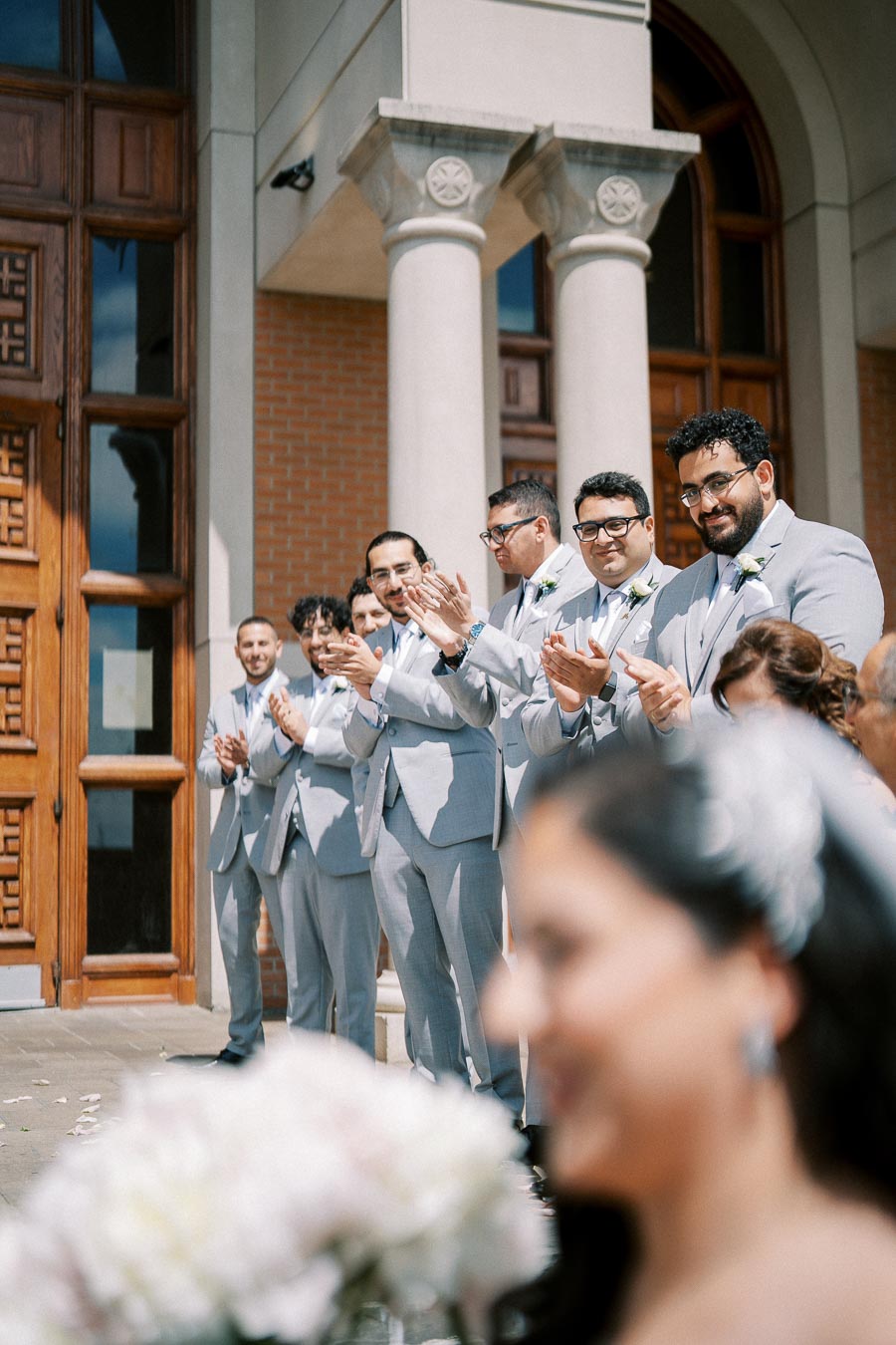 Groomsmen clapping in light gray suits at an outdoor wedding ceremony in front of a brick building with columns.