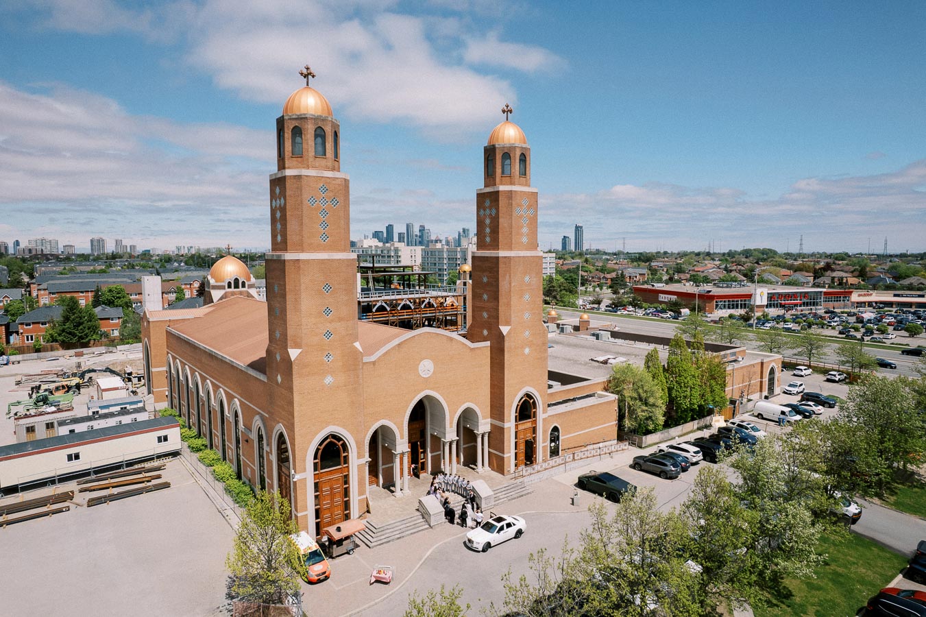 A large brick church with two copper-domed towers, set against a backdrop of a suburban landscape and city skyline, under a blue sky with scattered clouds.