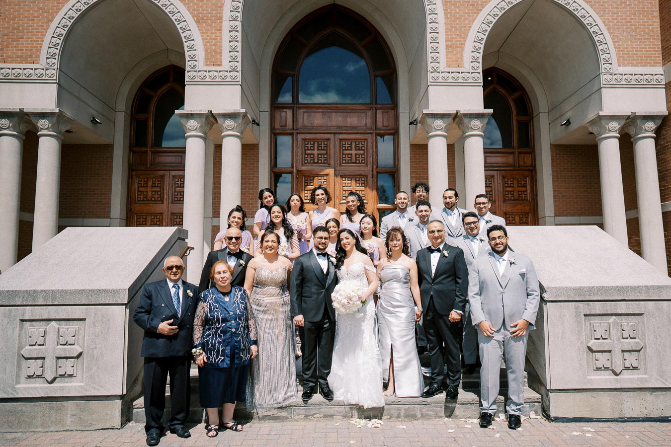 A large wedding party posing on the steps of an ornate building, featuring the bride and groom at the center surrounded by family and friends dressed in formal attire, with stone columns and detailed architectural elements in the background.