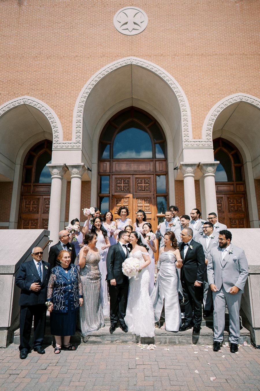 Wedding party gathered on the steps of a church, featuring a bride in a white gown and groom in a black tuxedo, surrounded by family and friends in formal attire, celebrating under a sunny sky with a brick building backdrop.