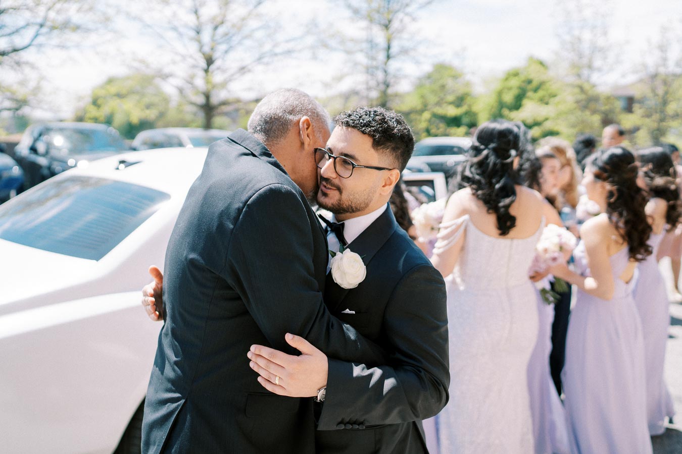 Wedding scene with groom in black suit and white rose boutonniere embracing an older man outdoors, while bridesmaids in lavender dresses gather in the background. A white car is parked nearby and trees are visible under the clear blue sky.