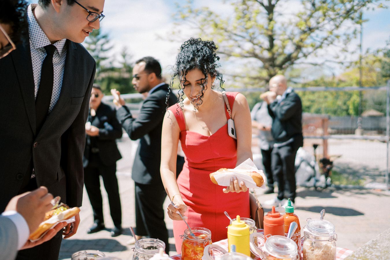 A woman in a red dress adds toppings to a hot dog at an outdoor gathering, surrounded by people in formal attire. Various condiments and jars are neatly arranged on a checkered tablecloth under a sunny sky.
