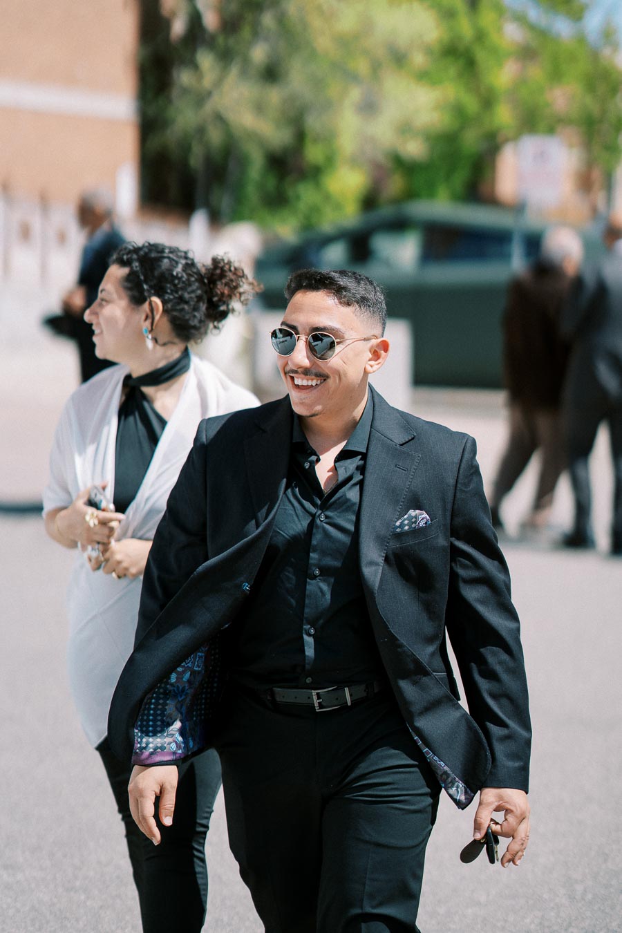 Two people walking outdoors in stylish attire; a man in a black suit and sunglasses smiling, accompanied by a woman in a white top and black pants.