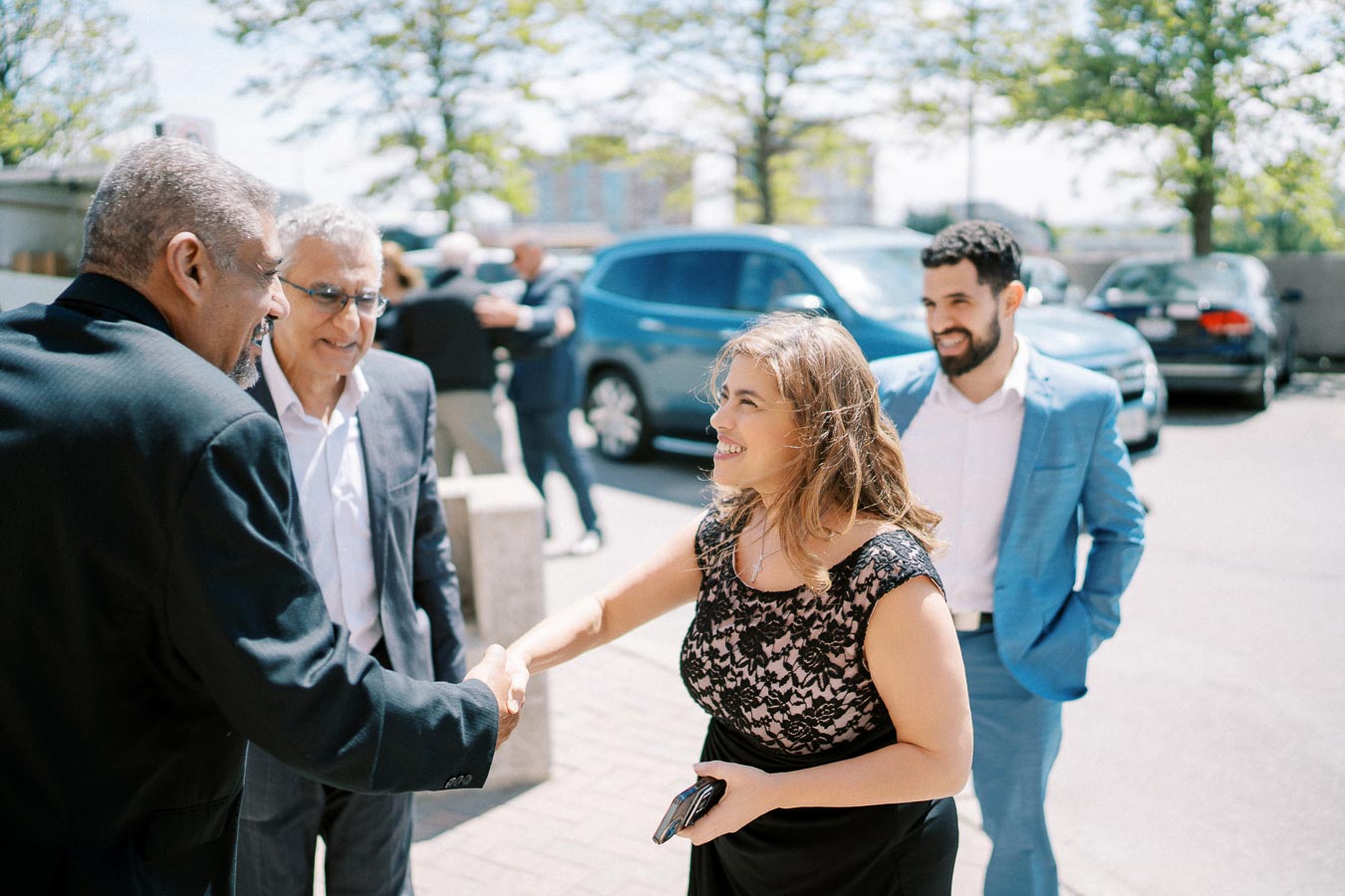 Group of business professionals shaking hands and smiling outdoors, with parked cars and trees in the background, illustrating a positive networking event or corporate meeting.