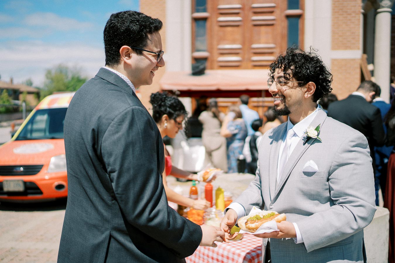 Two men in suits smiling and talking outdoors at a social gathering near a red food truck, with one man holding a hot dog. A woman in the background is serving condiments on a checkered table. The scene captures a celebratory and casual atmosphere.
