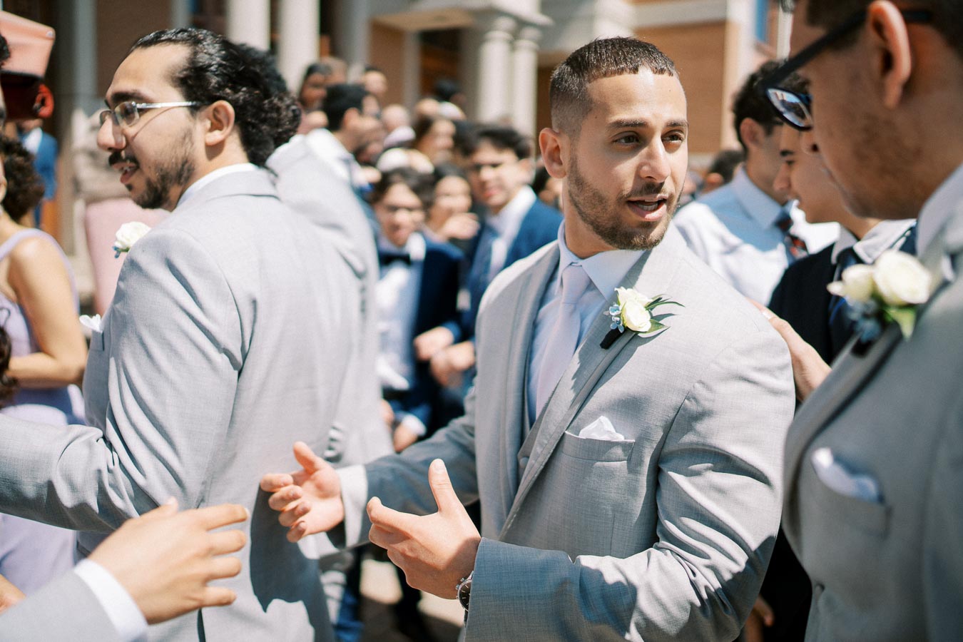 A group of men in light gray suits, some wearing boutonnières, engage in conversation at an outdoor wedding event, surrounded by other guests.