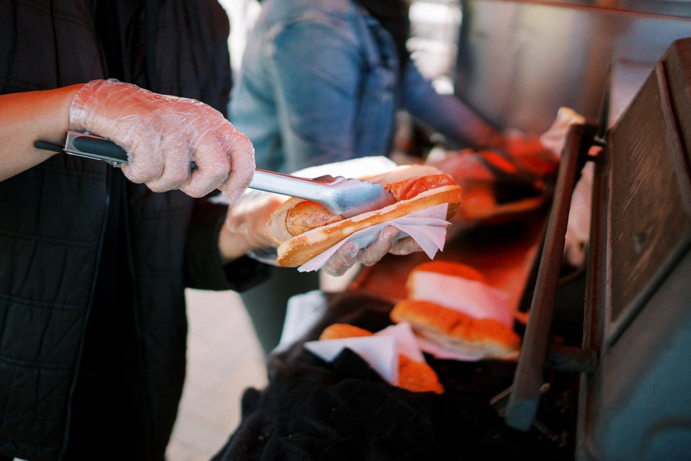 A person in disposable gloves uses tongs to prepare a fresh hot dog in a bun at a food stand.