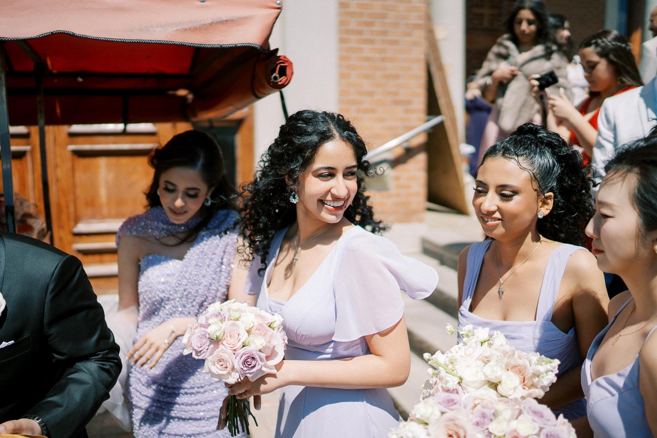 Bridesmaids in lavender dresses smiling and holding bouquets of pink and white roses at an outdoor wedding.