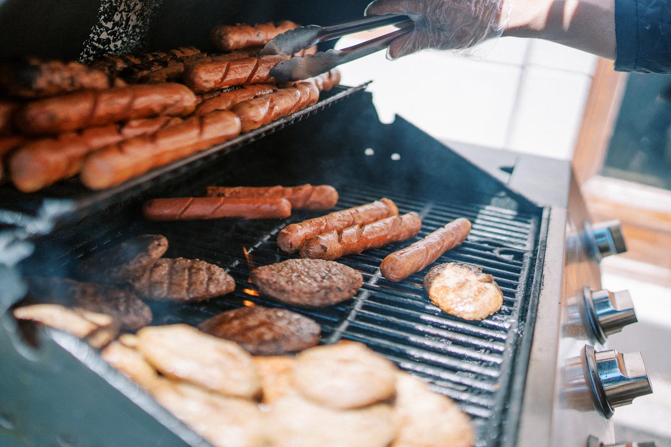 Grilling hot dogs and burgers on a barbecue with tongs, showcasing a summer outdoor cooking scene.