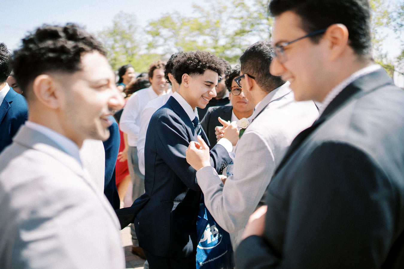 A group of men in suits having a friendly conversation at an outdoor event, with one man smiling and shaking hands, set against a background of trees and clear blue sky.