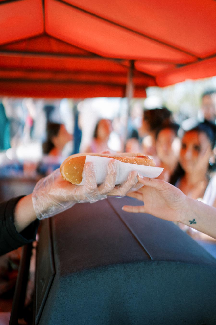 A vendor hands over a hot dog to a customer under a red canopy at an outdoor food stand, with a blurred crowd in the background at a sunny event.
