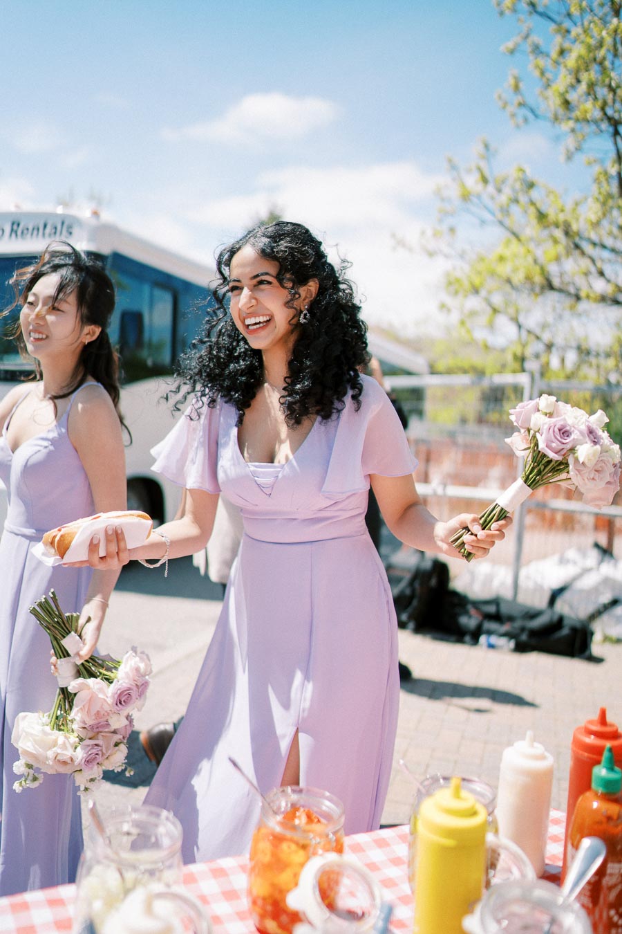 Bridesmaids in lavender dresses smiling and holding bouquets at an outdoor celebration, with condiments and a food truck in the background on a sunny day.
