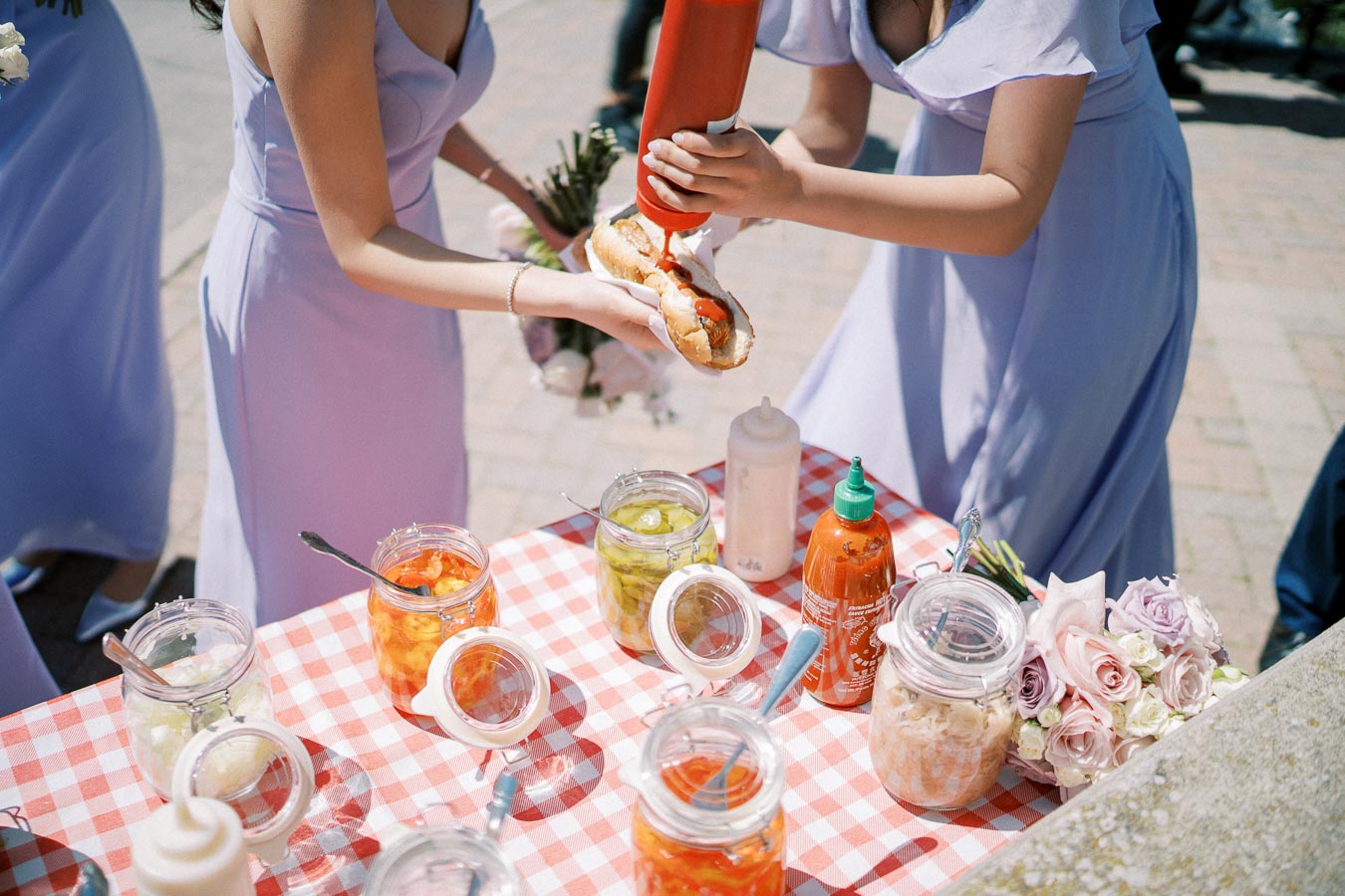 Two women in light purple dresses prepare a hot dog at an outdoor event, adding ketchup from a bottle. The table is covered with a red and white checkered tablecloth and has jars of various condiments and toppings. A bouquet of roses is visible on the side.