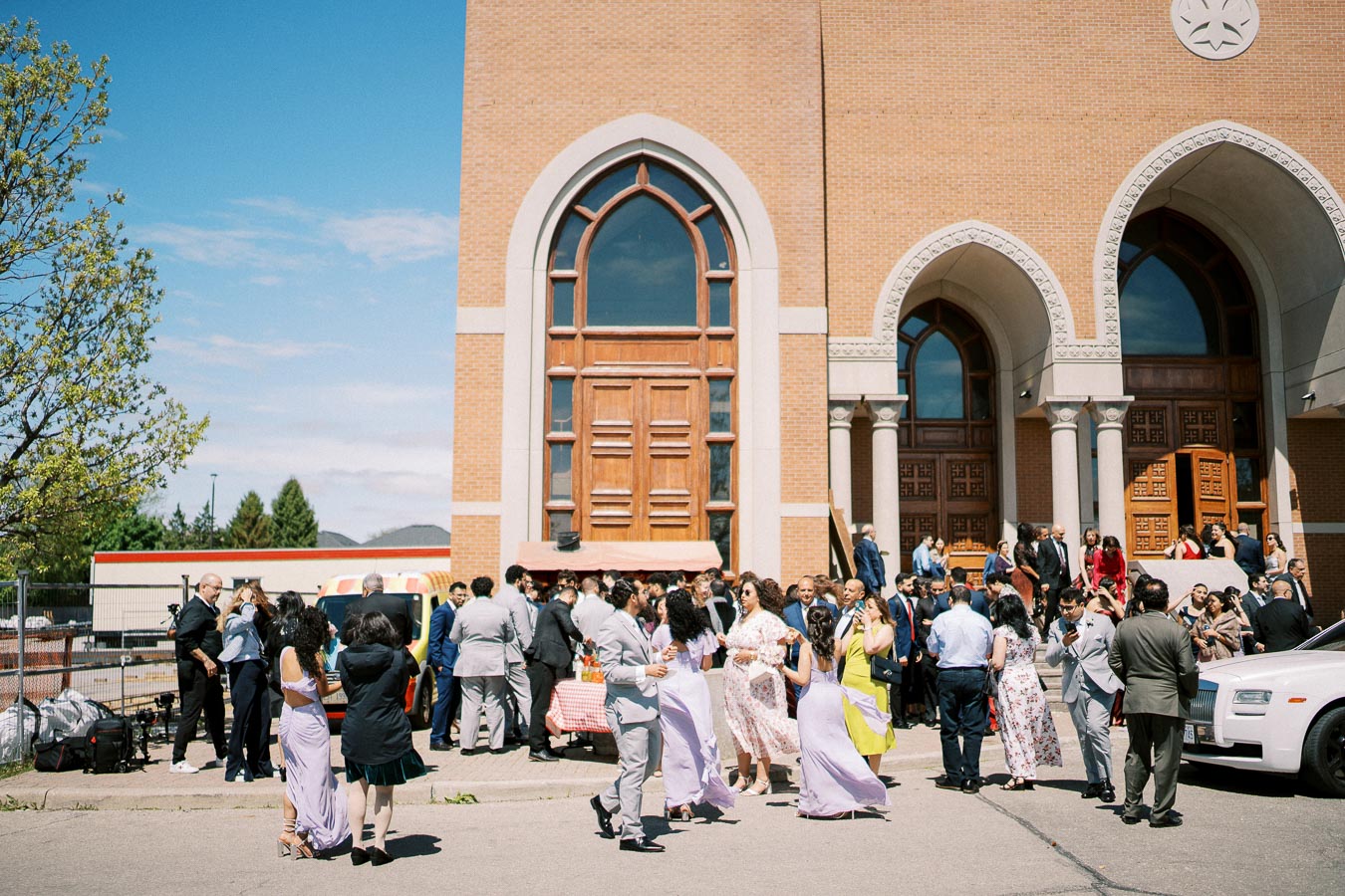 A large crowd of people in formal attire gathered outside a church with tall arched windows and wooden doors on a sunny day, possibly after a wedding ceremony.