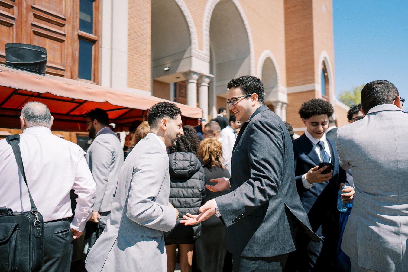 A group of well-dressed individuals engaging in conversation outside a building during a sunny day, with people smiling and interacting at a social event or gathering.