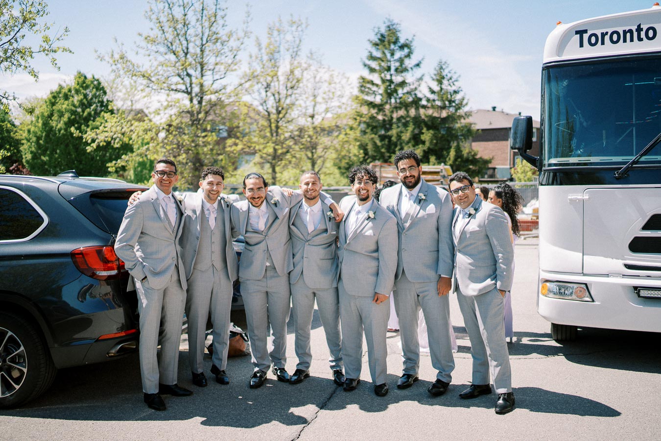 Group of groomsmen smiling in light gray suits with pink ties, standing near a bus labeled Toronto, with trees in the background, on a sunny day.