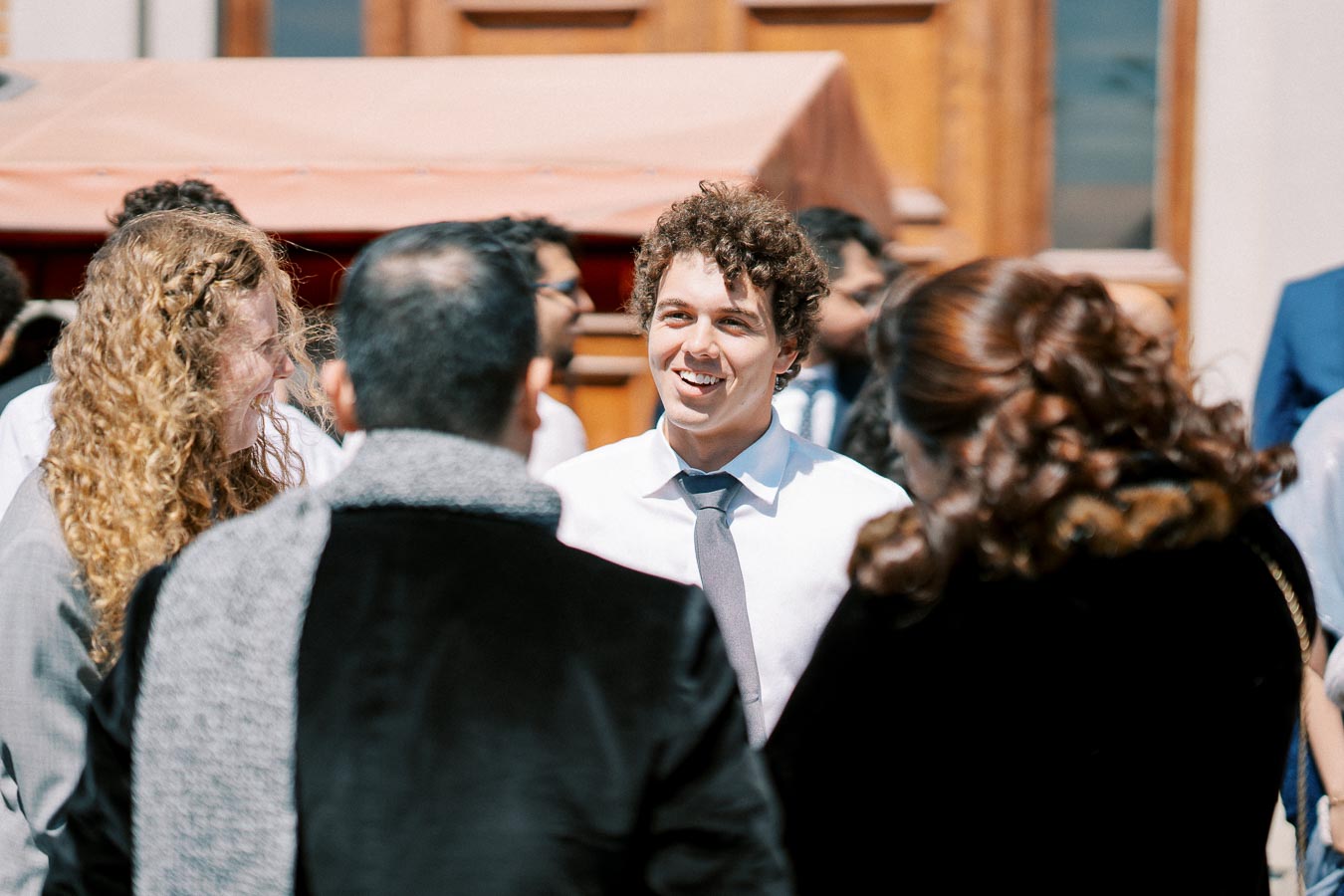 A group of people engaging in conversation at an outdoor event, with a smiling man in a white shirt and tie standing in the center, surrounded by others.