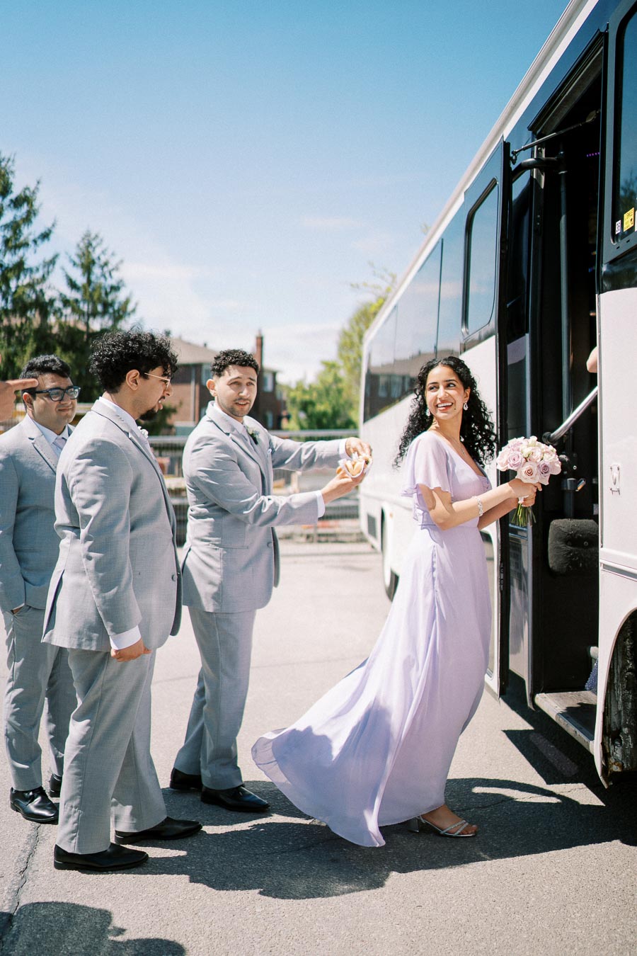 Bridesmaid boarding a bus holding a bouquet of light pink roses, accompanied by groomsmen in light gray suits on a sunny day.
