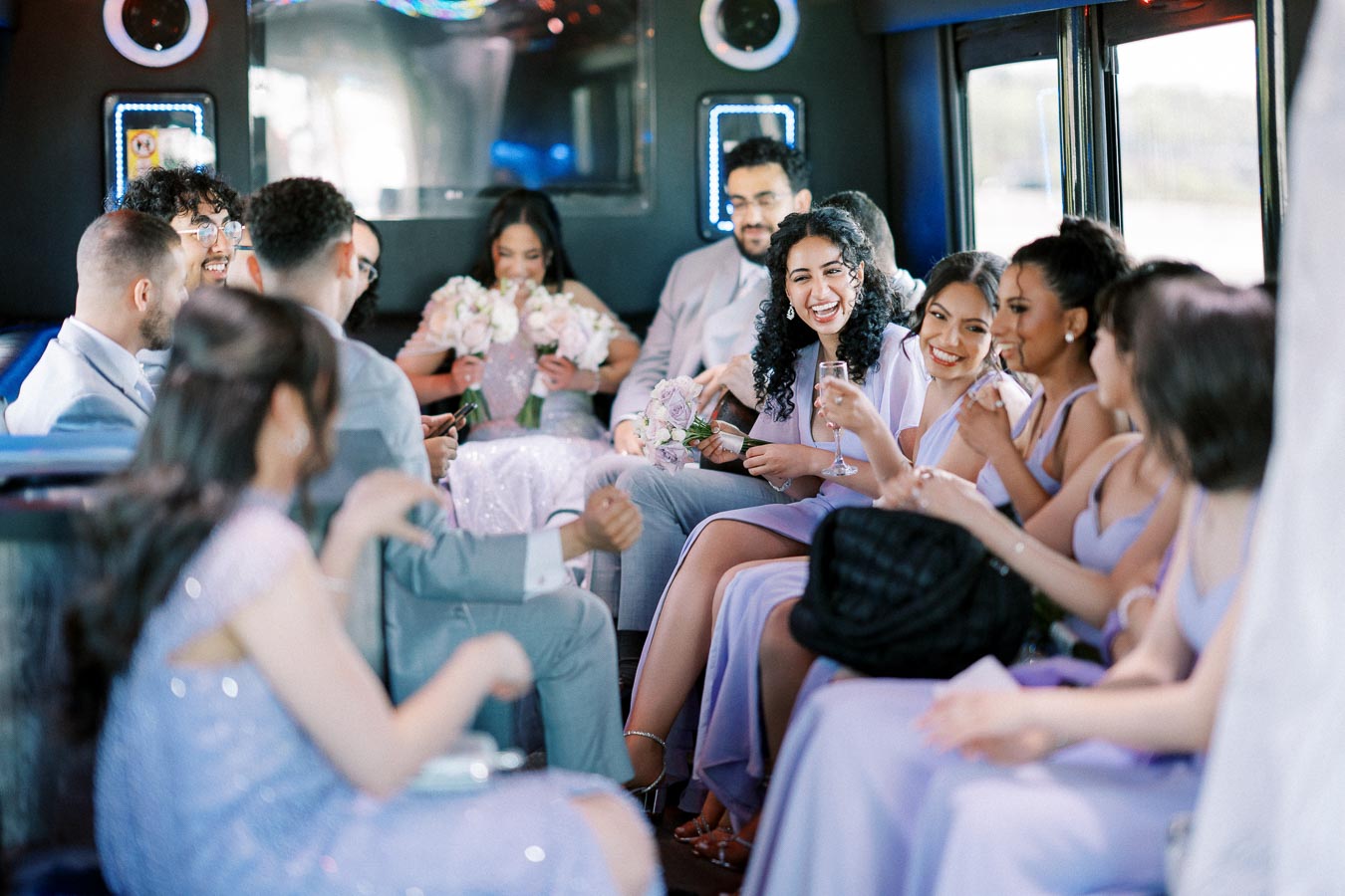 Group of people celebrating inside a party bus, wearing formal attire with pastel purple dresses and light-colored suits, holding bouquets and glasses, creating a joyful and festive atmosphere.