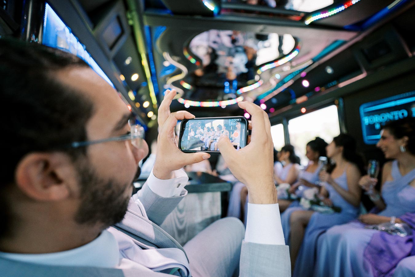 A man in a suit taking a photo with his smartphone inside a lively party bus with colorful lights, capturing a group of women dressed in elegant gowns.