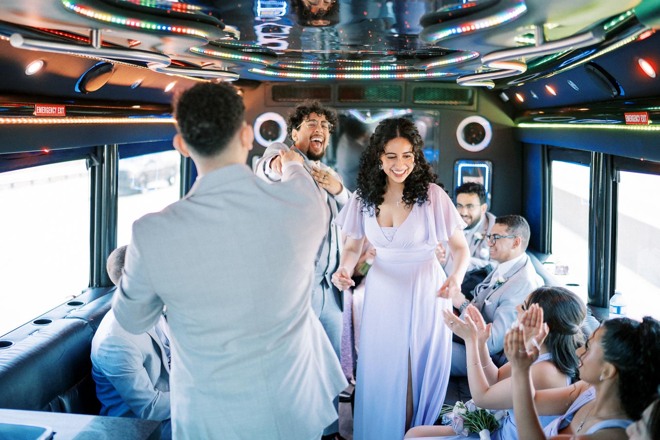 A lively group of elegantly dressed people dancing and celebrating inside a brightly lit party bus, featuring colorful LED lights reflecting off the ceiling.