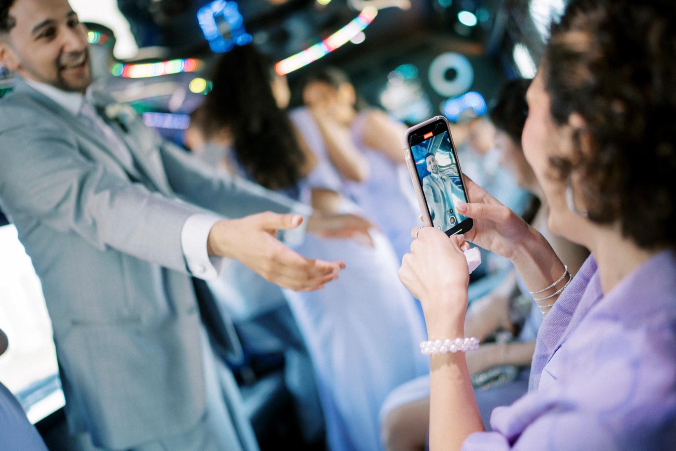A woman taking a photo with her smartphone in a party bus, focusing on a man in a light gray suit, with colorful lights in the background and guests wearing formal attire.
