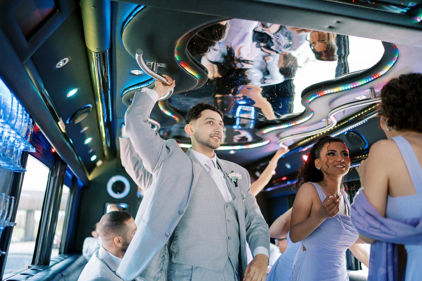 A group of elegantly dressed people enjoying themselves inside a vibrant, colorful limousine, featuring a futuristic ceiling with LED lights and reflections.