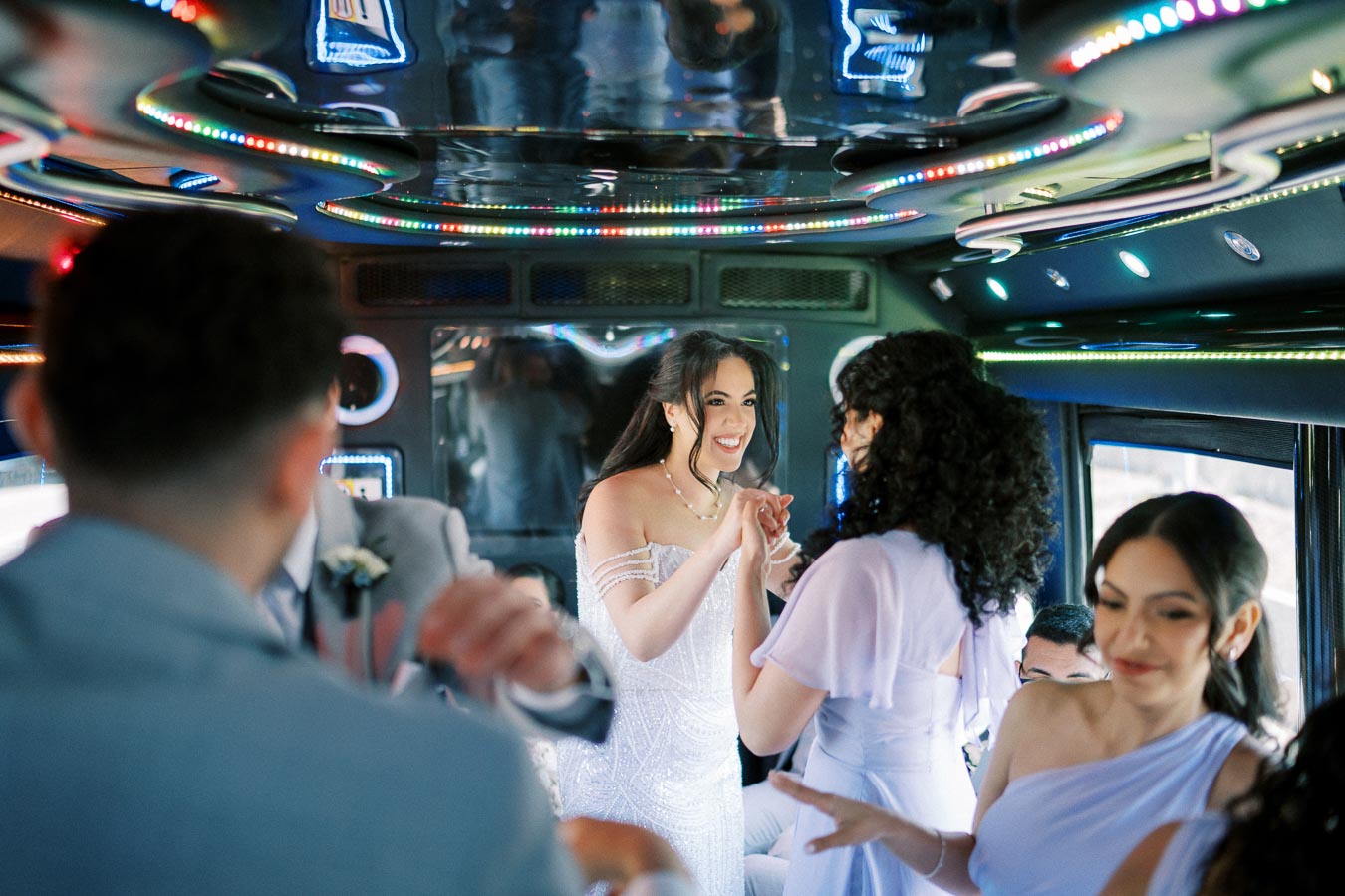 Group of people celebrating on a decorated party bus, featuring colorful interior lighting and stylish attire, with a focus on a smiling woman in a white dress.