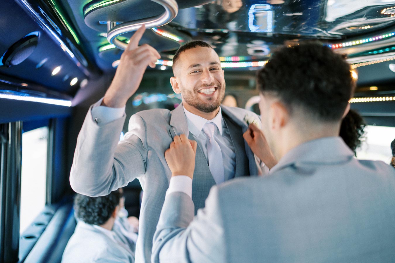 A smiling man in a light gray suit being helped with his jacket inside a festive, neon-lit party bus, surrounded by friends.