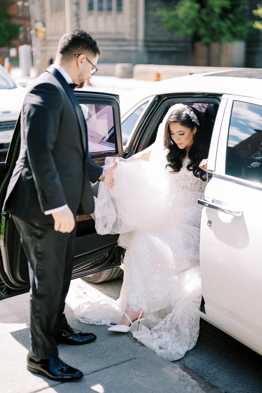 Bride elegantly exits white car with groom assisting, outdoor wedding scene.