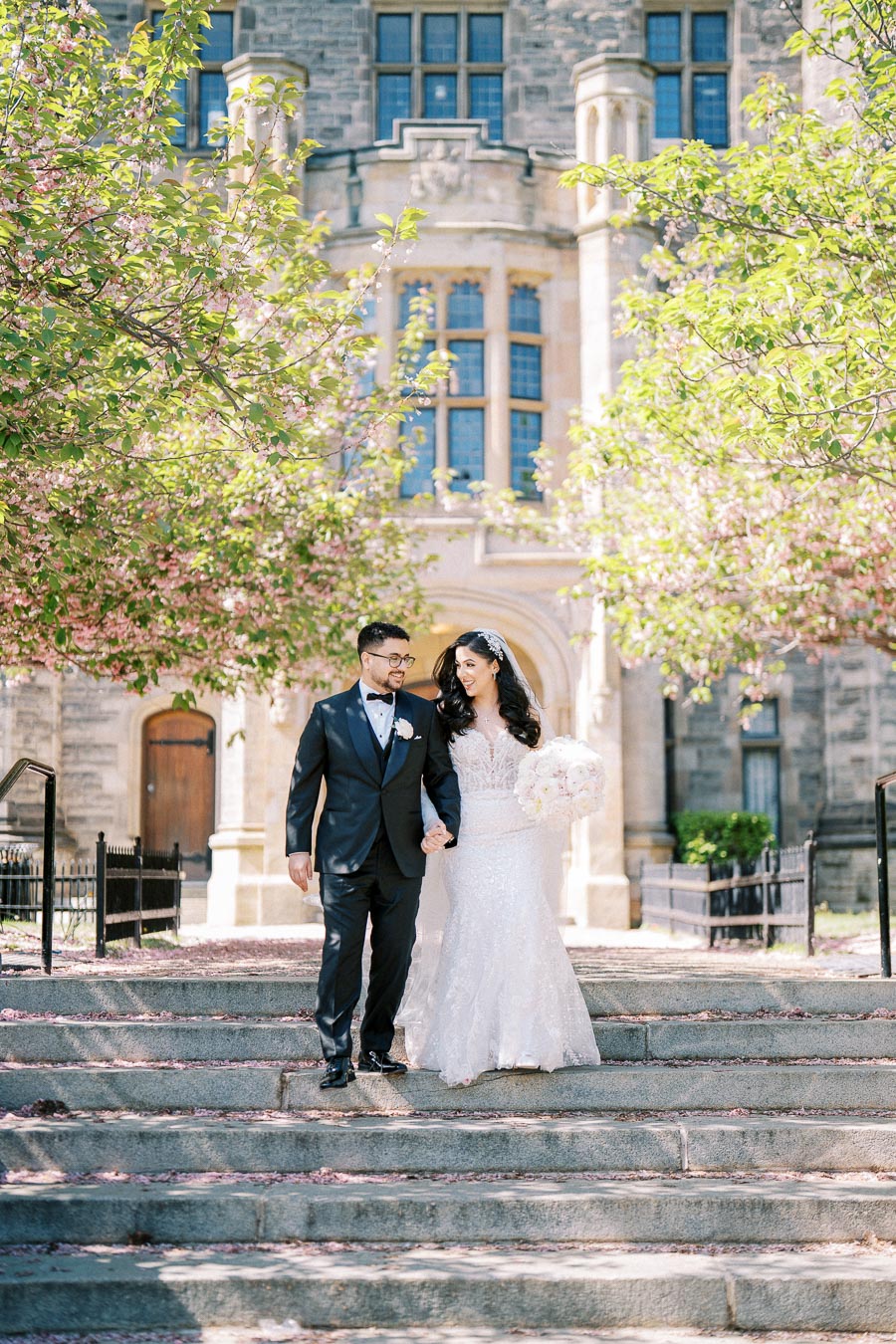 A newlywed couple in elegant wedding attire, walking hand in hand down stone steps surrounded by lush greenery and blooming trees, with a historic building in the background.