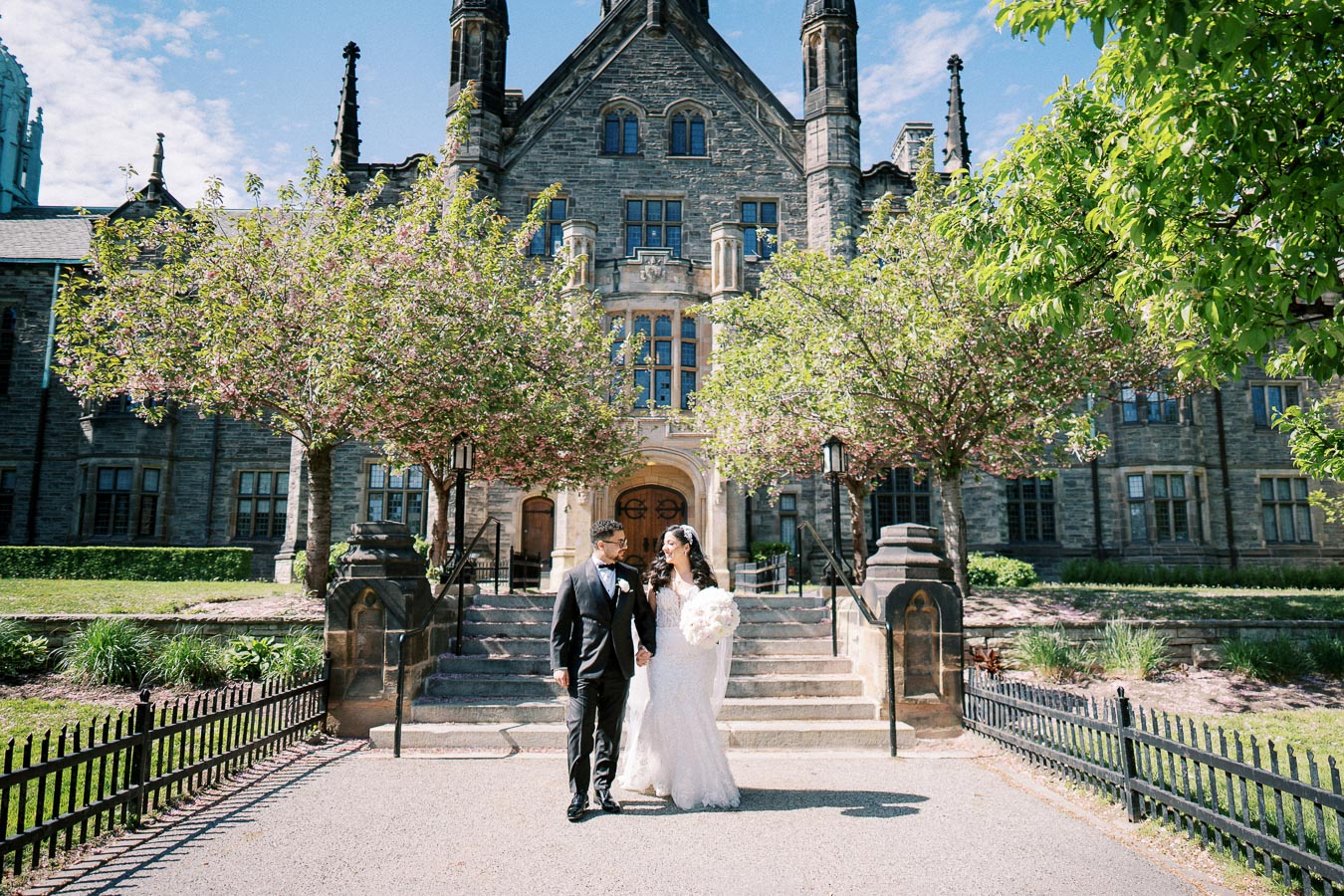 Wedding couple walking hand in hand in front of a historic stone building, surrounded by blooming spring trees, capturing a romantic moment on a sunny day.