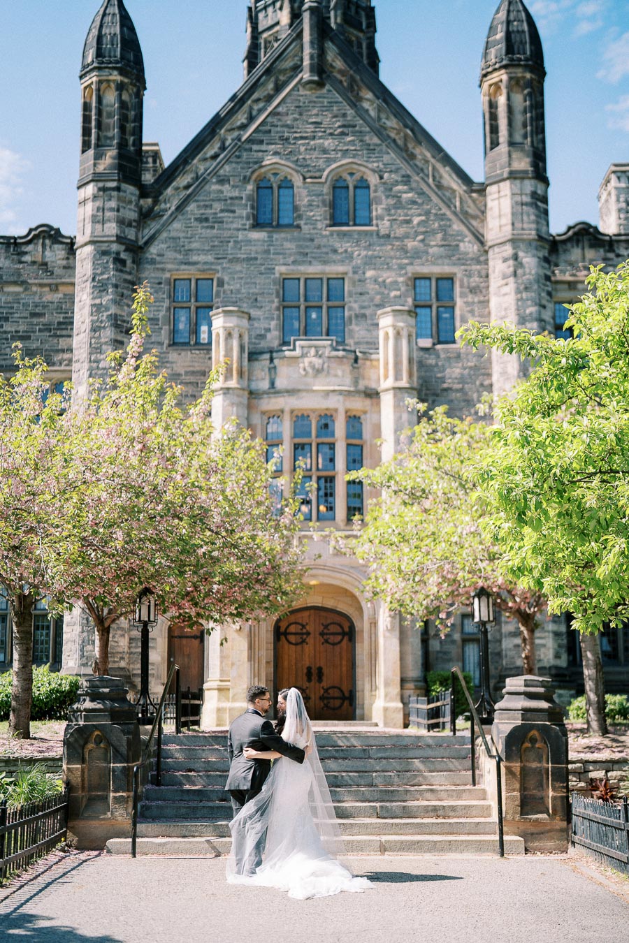 A couple in wedding attire stands in front of a historic stone building with arched windows, surrounded by blooming trees on a sunny day.