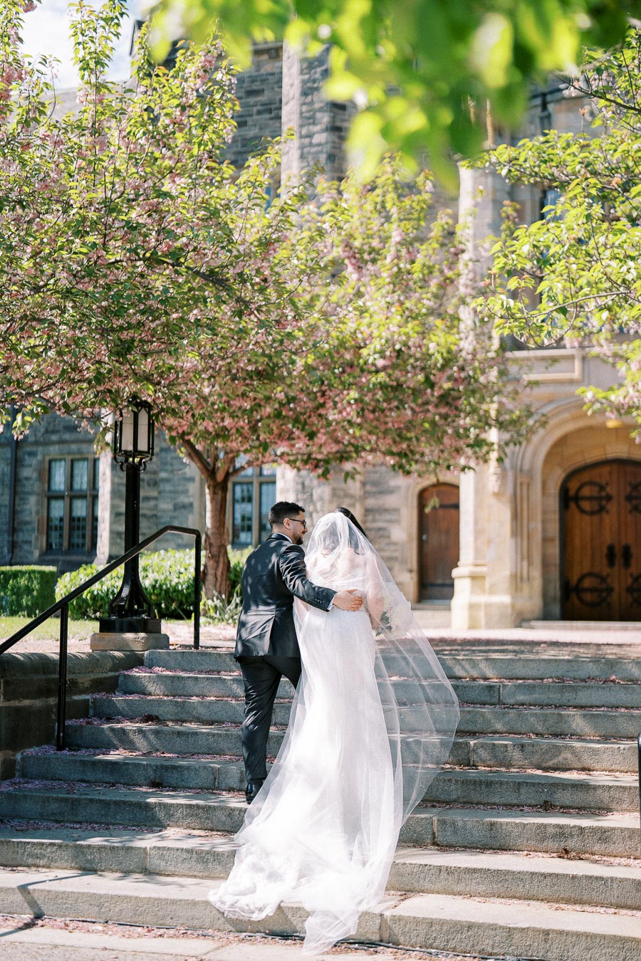 A bride and groom walking up stone steps towards an elegant building, surrounded by blooming trees, on a sunny day.